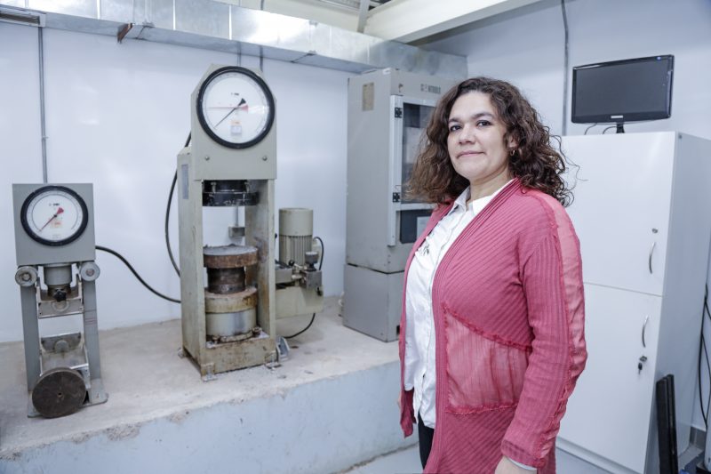 Woman standing in a laboratory/industrial testing room beside a hydraulic press and pressure gauges, smiling at the camera.