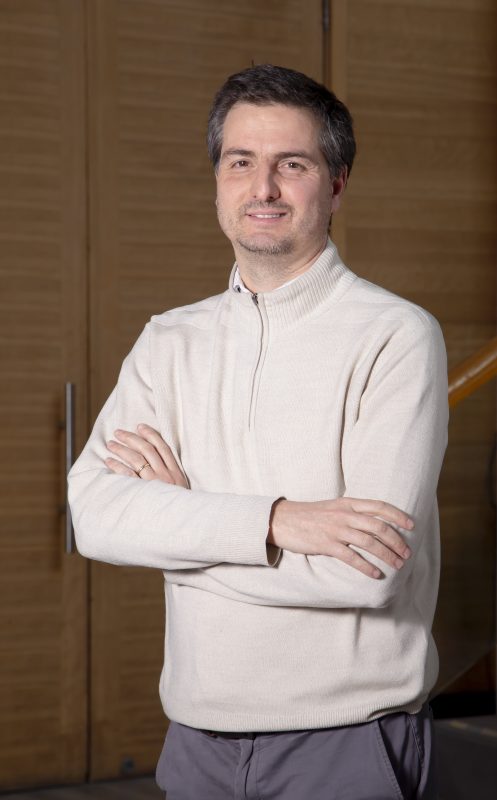Man wearing a light beige sweater stands with arms crossed, smiling, in a wood-paneled room.